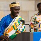 Marc wears a woven crown and bright blue tunic with a subtle floral pattern under a drape with tribal patterns in yellow, blue, green and white. He stands at a podium and holds a small Cote d'Ivoire flag.
