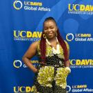 Nanyangwe Siuluta wears a black jumpsuit with large yellow and white geometric flowers and white dots, and a large white beaded necklace. Her hands are tucked into her pockets and she stands in front of a blue wall with repeating logos for UC Davis and Global Affairs in yellow and white. 
