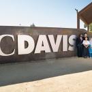 Eleven Humphrey fellows and two program coordinators stand next to a large UC Davis sign on campus. The sign is on a wall of gray concrete with large white letters that read "UC Davis". Humphrey fellows wear a mixture of business and traditional dress.