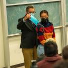 Keith David Watenpaugh stands at the front of a classroom putting a blue paper into a backpack worn by Naomi Danner. Both wear face coverings. Dr. Watenpaugh wears a dark green jacket and khaki pants, and dark rectangular framed glasses. His salt and pepper hair is neatly styled longer on top. Naomi stands in front of him wearing silver round-shaped framed glasses and a turtleneck blocked in navy blue, red, orange and navy blue, and light blue jeans. Her curly black hair is pulled back.