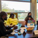 Gathered around a table with windows behind them looking out on trees and a grassy field, Edu Nguema, Rudo Chasi, a female student in a mask, Sushila Thing and a male student in a mask each have a laptop in front of them. Placards on the table identify this group as "Developing B Nations" and some of the countries they represent as Fiji and Nepal. 