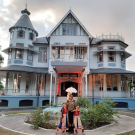 Kara Roopsingh stands in front of a light blue two-story wooden French provincial-style house with a wrap-around porch and frilly cast iron railings. With Kara are two little girls dressed identically and smiling with her. 