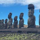Row of famous statues on Easter Island with hills in the background and a grassy stone foreground. 