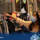 Two students wearing face masks stand in front of a rack of clothing and one points to a piece of art on the wall over the racks with a 19th century American or European mother and child. At the bottom of the image is a blue overlay with the Global Affairs logo on the left and the "Global Aggies" logo and iconography on the right.