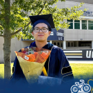 Jeff wearing a graduation cap and gown, holding flowers, and standing in front of a UC Davis sign.
