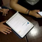Hands holding digital devices with the Article 26 Backpack website on the screen