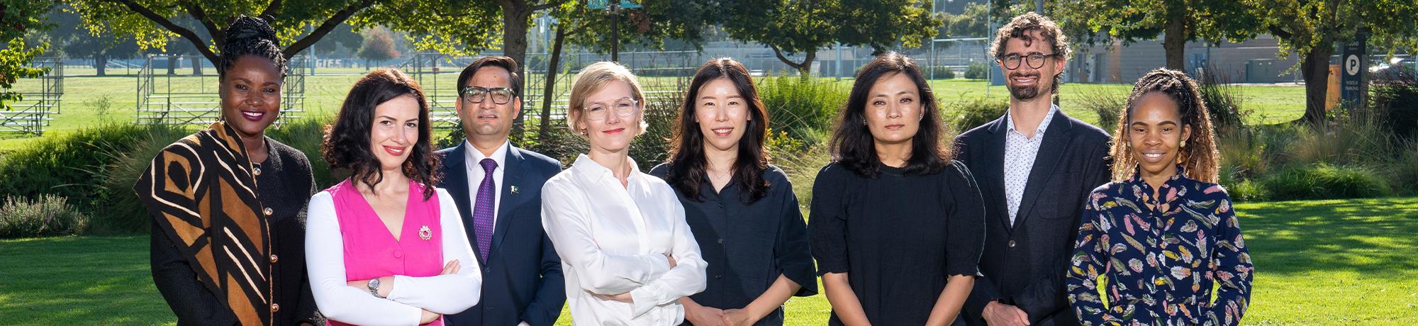 Eight Humphrey Fellows stand side by side smiling at the camera with trees and shrubbery behind them in the background.