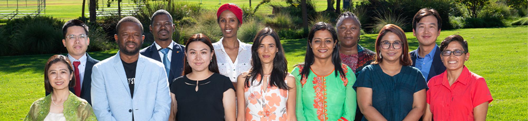 Twelve Humphrey Fellows stand together in two rows on the grass of a lawn on the UC Davis campus in the sunlight. They wear a combination of culturally traditional and business dress. 