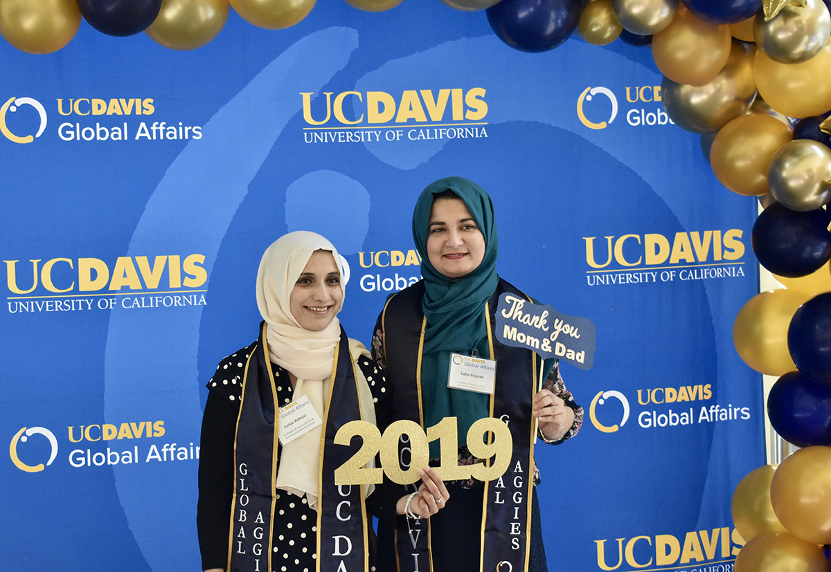 two women in hijab standing in front of a blue background with balloons holding a sign that says 2019